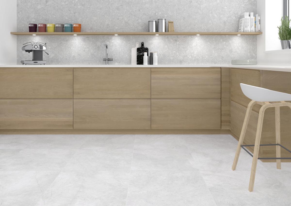 Eine moderne L-Küche mit grifflosen Fronten aus hellem Eichenholz, einer weißen Arbeitsplatte und hellgrauen Bodenfliesen in Naturstein-Optik. Die Küchenrückwand zeigt sich ebenfalls in grauem Naturstein-Design, ergänzt durch ein langes Holzregal mit integrierter LED-Beleuchtung und Küchenutensilien.