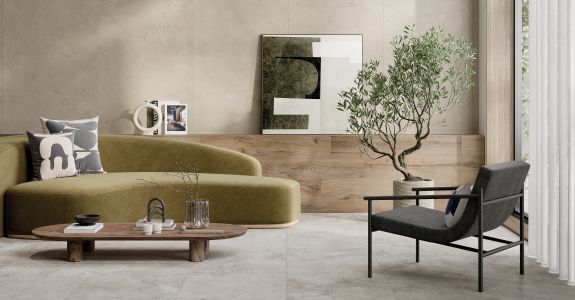 Ein stilvoll eingerichtetes, modernes Wohnzimmer mit einem olivgrünen Sofa. Davor steht ein ovaler Couchtisch aus Holz. Rechts im Bild befindet sich ein schwarzer Sessel. Die Wand ist teils mit beige-grauen Steinfliesen, der Boden mit oßformatigen, hellgrauen Feinsteinzeugfliesen ausgelegt.