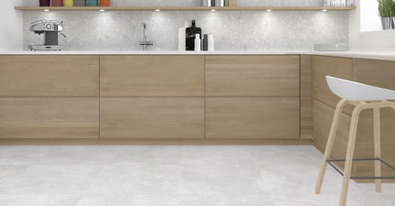 Eine moderne L-Küche mit grifflosen Fronten aus hellem Eichenholz, einer weißen Arbeitsplatte und hellgrauen Bodenfliesen in Naturstein-Optik. Die Küchenrückwand zeigt sich ebenfalls in grauem Naturstein-Design, ergänzt durch ein langes Holzregal mit integrierter LED-Beleuchtung und Küchenutensilien.