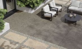 Moderne Outdoor-Terrasse mit großformatigen, quadratischen Feinsteinzeugfliesen in natürlichem Grauton. Die Fliesen haben eine feine, steinähnliche Struktur und sind im Format 60 x 60 cm verlegt. Sie wirken robust, rutschfest und sind ideal für den Außenbereich geeignet. Im Hintergrund ist eine gemütliche Sitzgruppe mit hellem Polster und ein runder, schwarzer Tisch zu sehen, eingerahmt von grünen Pflanzen und beige-grauen Fliesen im Kiesbett.