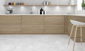 Eine moderne L-Küche mit grifflosen Fronten aus hellem Eichenholz, einer weißen Arbeitsplatte und hellgrauen Bodenfliesen in Naturstein-Optik. Die Küchenrückwand zeigt sich ebenfalls in grauem Naturstein-Design, ergänzt durch ein langes Holzregal mit integrierter LED-Beleuchtung und Küchenutensilien.